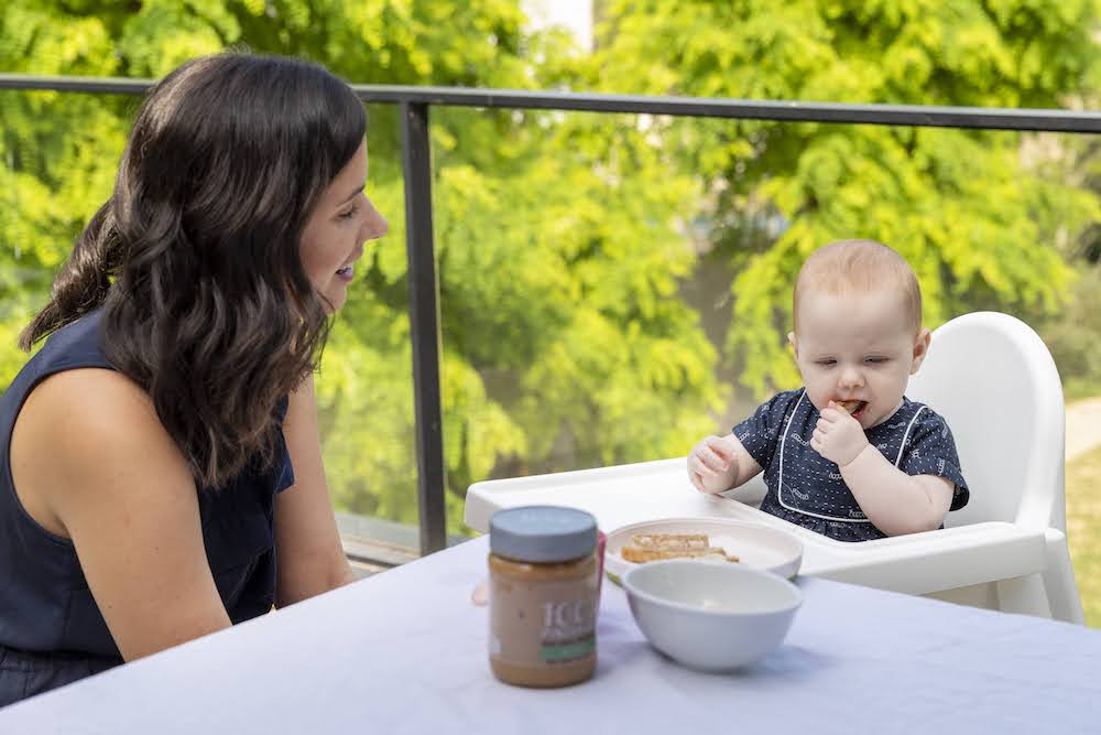 mama zit buiten aan tafel met baby die boterhammen met pindakaas eet 